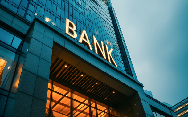 A modern tall building in dark blue with a bank sign at the entrance, featuring a bright yellow light and an exterior fully covered in windows.